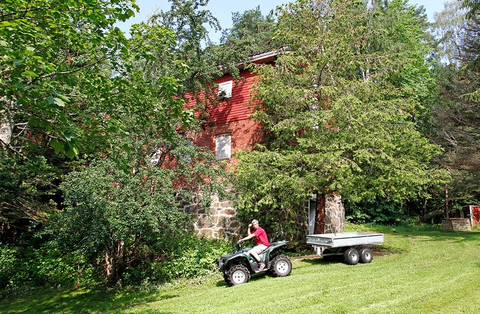 En person kjører en ATV med tilhenger på en gressplen ved siden av en rød, toetasjes bygning i stein og tre omkranset av trær.